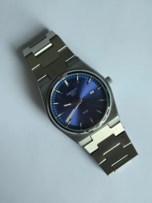 Tissot PRX klocka med blå urtavla - Tissot armbandsur för unisex med blå urtavla och datumvisning. Boett och länk i rostfritt stål med viklås. Analog visning med tim-, minut- och sekundvisare. Passar som vardagsklocka eller för mer formella tillfällen.  Nypris 4000-5000sek Vårat mycket billigare pris: 2999:-  Obs... klockan har en liten skråma på baksidan då det ser ut som att någon har råkat rista upp den när man försökt att öppna Baksidan, därav det billiga priset