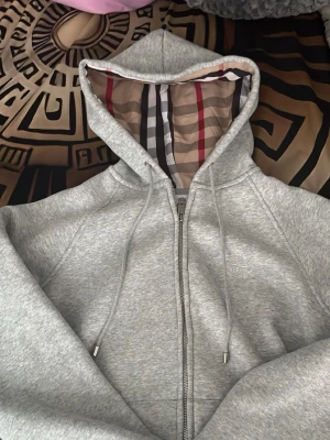Grå zip-hoodie från Burberry - Snygg grå hoodie med dragkedja från Burberry. Hoodien har klassiskt rutigt foder i huvan, dragsko och diskret broderad logga på bröstet. Perfekt för en stilren och trendig look. Mjuk bomullsblandning för extra komfort.