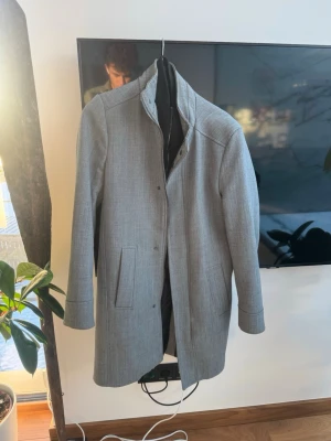 Ljusgrå klassisk kappa - Stilren ljusgrå kappa i rak modell med hög krage och dolda knappar. Två sidofickor och diskret struktur i tyget ger en modern look. Perfekt för dig som vill ha en enkel men snygg ytterrock till hösten eller vintern.