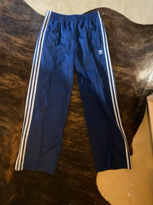 Blå Adidas mjukisbyxor med vita ränder - Säljer ett par klassiska blå Adidas mjukisbyxor med vita ränder längs benen och broderad logga på vänster sida. Byxorna har resår i midjan och dragkedjefickor. Perfekta för chill eller streetwear-stil.