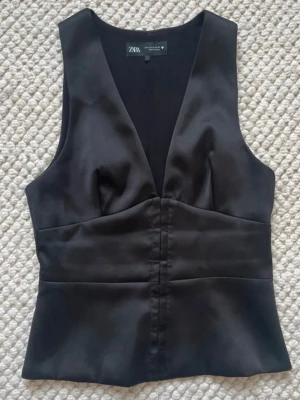 Svart v-ringad väst från Zara - Stilren svart väst från Zara med v-ringning och figurnära passform. Västen har slät yta och bred midjedel som ger en snygg siluett. Perfekt att styla över en skjorta eller t-shirt för en trendig look.