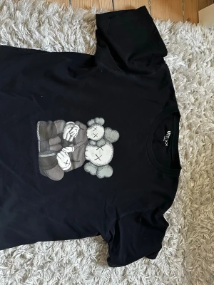 Kaws T-Shirt - Säljer en svart t-shirt från Uniqlo collab med KAWS. Tröjan har ett stort KAWS-motiv på framsidan och två X på ryggen. T-shirten har rund hals och korta ärmar. Perfekt för dig som minns streetwear och gillar coola prints.