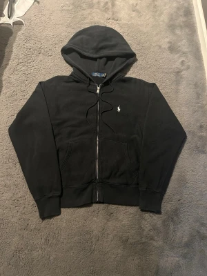 Svart hoodie från Polo Ralph Lauren - Klassisk svart hoodie med dragkedja från Polo Ralph Lauren. Har huva med dragsko, två fickor framtill och den ikoniska vita loggan broderad på bröstet. Tillverkad i mjukt bomullsmaterial som är skönt att bära.