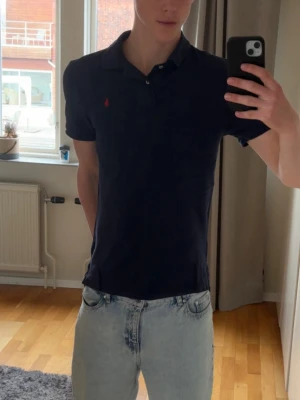 Mörkblå pikétröja från Polo Ralph Lauren - Säljer en klassisk mörkblå pikétröja från Polo Ralph Lauren med korta ärmar och krage. Tröjan har en liten röd broderad logga på bröstet och är tillverkad i bomull. Perfekt för en avslappnad och stilren look. Perfekt nu till sommaren. Skick 9/10