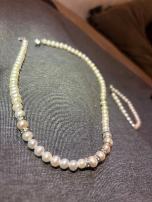 Pärlhalsband 18k - Säljer ett elegant  set halsband och armband med vita pärlor och små silver cross-over detaljer mellan vissa pärlor. Halsbandet har ett dekor visible lås i silverfärg. Perfekt för dig som gillar klassisk och stilren accessoar med en modern twist.