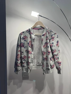 Grå hoodie med rosa blommor - Säljer en grå hoodie med dragkedja och rosa blommönster över hela plagget. Tröjan har ribbade muddar vid ärmslut och nederkant samt två fickor framtill. Perfekt för dig som gillar en söt och avslappnad stil.