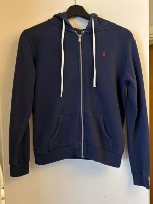 Mörkblå hoodie från Polo Ralph Lauren - Mörkblå hoodie med dragkedja från Polo Ralph Lauren. Klassisk design med vit dragsko i huvan, två fickor framtill och röd broderad logga på bröstet. Tillverkad i mjukt bomullsmaterial som är perfekt för en avslappnad stil. Nypris 2995kr. 