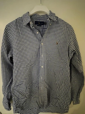 Rutig soprtig skjorta från Ralph soprtig - Klassisk rutig skjorta från Ralph Lauren i blått och vitt soprtigt mönster. Skjortan har lång ärm, knappar framtill och en liten broderad logga på bröstet. Tillverkad i mjuk bomull för en soprtig och bekväm känsla.
