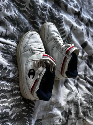 Vita Champion sneakers med röd detalj - Snygga vita sneakers från Champion med röda och marinblå detaljer. Skorna har perforerade sidor, klassisk snörning och högt skaft. Tillverkade i läderimitation med bekväm vadderad insida och Champion-logga på sidan och plösen. Använd 2-3 gånger.