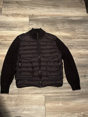 Moncler cardigan  - Svart cardiganjacka med quiltad, vadderad front och stickade ärmar. Jackan har dragkedja framtill, två fickor med dragkedja och ribbad krage. Perfekt för lager-på-lager och en clean look.