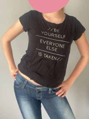 Svart t-shirt med texttryck - Svart kortärmad t-shirt med vitt texttryck på framsidan där det står 'Be yourself, everyone else is taken'. T-shirten har en rund halsringning och en avslappnad passform. Perfekt till jeans för en chill look.