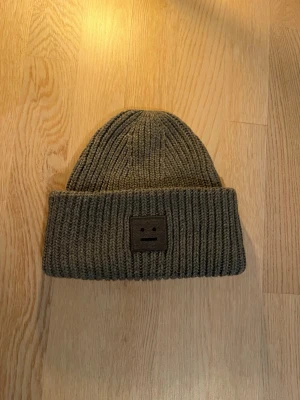 Grå mössa från Acne Studios - Säljer en ribbstickad olivgrön mössa från Acne Studios med uppvikt kant och det klassiska ansiktslogotypen framtill. Mössan är gjord i mjukt, tjockt material som håller värmen och har en clean, minimalistisk look som passar till det mesta.