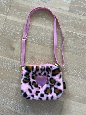 Rosa axelväska med leopardmönster - Säljer en fluffig axelväska i rosa med leopardmönster och ett lila hjärta i mitten. Väskan har justerbar axelrem i rosa konstläder och dragkedja upptill. Perfekt för dig som gillar statement-accessoarer och djurmönster.