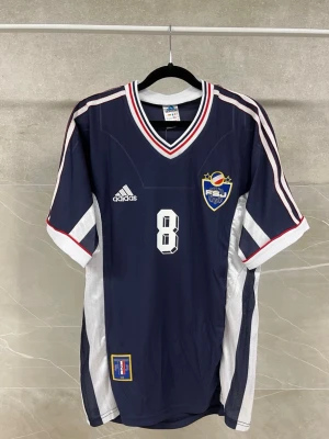 Adidas fotbollströja Mijatovic #8 - Mörkblå fotbollströja från Adidas med vita och röda detaljer, nummer 8 och namnet Mijatovic på ryggen. Tröjan har klubbmärke på bröstet, V-ringad krage och korta ärmar. Perfekt för fotbollsfans som gillar retrostil.  Finns i olika storlekar!!! Pris kan diskuteras!!!