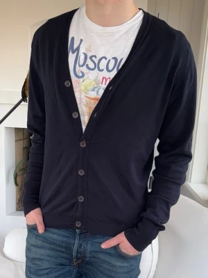 Svart basic kofta med knappar - En enkel svart kofta med långa ärmar och knäppning framtill. Koftan har en smal passform och ribbade muddar vid ärmslut och nederkant. Perfekt att slänga över en t-shirt för en avslappnad look.