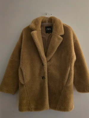 Beige teddyjacka från Zara - Mysig teddyjacka från Zara i beige färg med klassisk krage och två knappar framtill. Jackan har en rak och något oversize passform, samt två sidofickor. Materialet är fluffigt och mjukt, perfekt för att hålla värmen under kalla dagar.