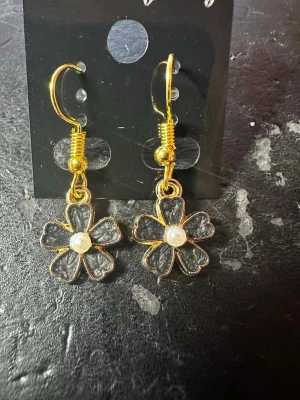 Blommiga örhängen med pärldetalj - Snygga örhängen i form av blommor med guldfärgade kanter och svart fyllning. I mitten av varje blomma sitter en liten vit pärldetalj som ger extra glans. Perfekta för dig som vill ha en söt och trendig accessoar till din outfit.