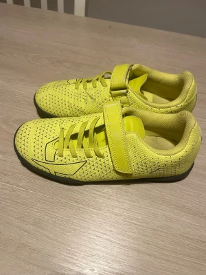 Gula fotbollsskor med kardborreband - Snygga gula fotbollsskor med svart mönster och kardborre clousure för enkel på”; och avtagning. Skorna har snörning framt complete fit och en låg profil direct för inomhusspel. Ovandelen är i syntetmaterial som ger bra bollkontroll.