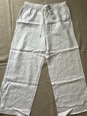 Mng Mango Vita linnebyxor med dragsko - Säljer ett par fräscha vita linnebyxor från Mango  med avslappnad passform och elastisk midja med dragsko. Byxorna har raka ben och är tillverkade i luftigt linne, perfekta för en chill och stilren look.