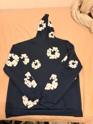 Blå hoodie från Denim Tears med tryck - Snygg mörkblå hoodie från Denim Tears med vita blomliknande tryck över hela tröjan och huvan. Hoodien har en klassisk passform, huva och känguruficka framtill. Perfekt för dig som vill sticka ut med en unik streetwear-look. Mer frågor så är det bara och skriva!