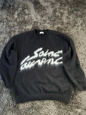 Svart Saint Laurent stickad tröja - Stickad svart tröja från Saint Laurent med vit fuzzy logga framtill. Tröjan har rund halsringning, långa ärmar och ribbade muddar. Materialet ser ut att vara mohairmix vilket ger en fluffig och mjuk känsla. Perfekt för dig som gillar streetstyle och exklusiva märken.