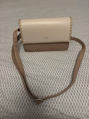 Beige axelväska från Ulrika - Snygg axelväska från Ulrika i två nyanser av beige med dekorativa sömmar längs kanten. Väskan har ett lock med magnetknäppning och justerbar axelrem. Insidan har fack med dragkedja för extra förvaring. Perfekt för att bära dina viktigaste saker.