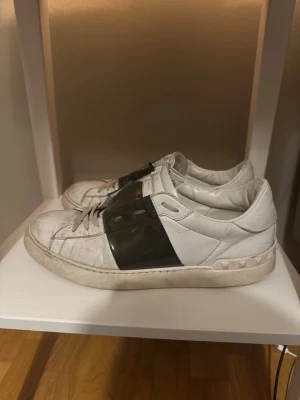 Vita sneakers från Valentino 40 - Säljer ett par vita sneakers från Valentino med en bred svart rem över mitten och coola nitar runt hälen. Skorna är i läder och har en chunky sula som ger en modern vibe. Perfekta för dig som vill sticka ut med en lyxig touch.
