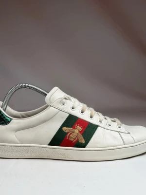 Gucci Ace vita sneakers med bi - Gucci Ace sneakers i vitt läder med klassiska gröna och röda sidoremsor och broderad guldfärgad bi. Skon har vita snören och grön detalj på spännet bak. Snygg och tidlös modell som sticker ut med sina exklusiva detaljer.
