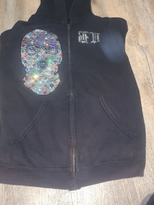 Svart hoodie med strassmask och tryck - Svart zip-hoodie med stor ficka framtill, dekorerad med en färgglad strassapplikation i form av en mask på vänster sida och ett vitt tryck med bokstäverna 'EW' på bröstet. Mjuk bomullskänsla och avslappnad passform, perfekt för en streetwear-look.