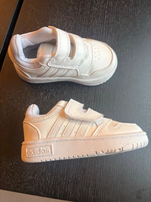 Vita sneakers med kardborre från Adidas - Säljer ett par vita sneakers från Adidas med klassiska tre ränder på sidan och kardborreband för enkel påtagning. Skorna har en låg profil och är tillverkade i syntetmaterial med vit sula och meshfoder för extra komfort.