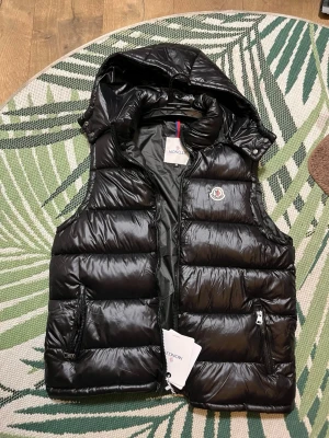 Svart dunväst från Moncler med huva - Storlek M passar S aldrig annvänd och bara teatad Snygg svart dunväst från Moncler med glansig finish, avtagbar huva och dragkedja framtill. Västen har två fickor med dragkedja och klassisk Moncler-logga på bröstet. Insidan har en serietidningsdetalj och märkestaggar.