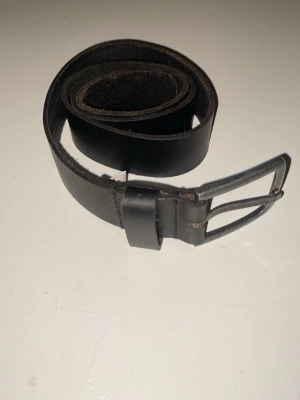 Svart klassiskt skärp i skinn - Snyggt och stilrent svart bälte i äkta skinn med enkel metallspänne. Perfekt till jeans eller byxor för en clean look. Passar dig som gillar minimalistisk stil och vill ha ett bälte som funkar till det mesta.