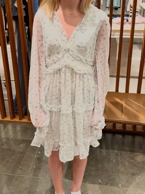 Vit blommig omlottklänning med volang - Romantisk vit kortklänning med små blommor, omlott-design och volangdetaljer. Klänningen har långärmade transparenta ärmar och v-ringad hals. Perfekt för sommarens alla tillfällen. 