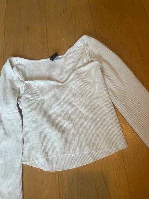 Vit ribbad offshoulder topp - Trendig vit ribbad topp med offshoulder-design och långa ärmar. Toppen har en croppad passform och är tillverkad i ett mjukt, stretchigt material som sitter snyggt på kroppen. Perfekt för dig som vill ha en stilren och modern look.