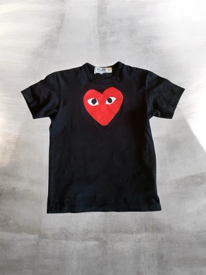 Svart Comme des Garçons PLAY t-shirt - Säljer en svart t-shirt från Comme des Garçons PLAY med det ikoniska röda hjärtat med ögon på bröstet. T-shirten är i 100% bomull och har en klassisk rak passform och rund hals. I riktigt bra skick. Om du har några frågor eller funderingar är det bara att höra av dig. Priset är ej hugget i sten!