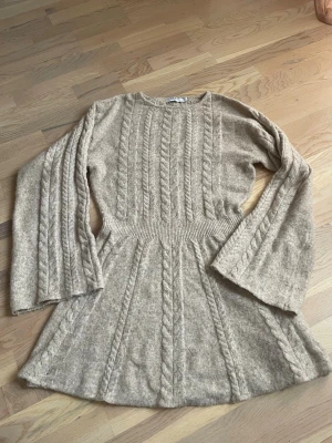 Beige stickad klänning från NA-KD - Mysig beige stickad klänning från NA-KD med kabelstickat mönster och rund hals. Klänningen har långa, vida ärmar och markerad midja för en snygg siluett. Perfekt för kyligare dagar och enkel att styla med boots och ett par strumpbyxor! 