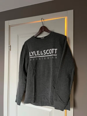 Grå sweatshirt från Lyle & Scott - Mörkgrå sweatshirt med vit Lyle & Scott Sportswear-logga på bröstet. Tröjan har rund halsringning, långa ärmar och ribbade muddar. Perfekt för en avslappnad och sportig stil.