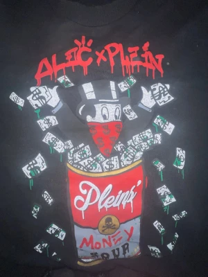 Philipp&Plein svart hoodie med tryck - Svart hoodie från Philipp Plein x Alec med stort färgstarkt tryck framtill som visar en tecknad figur med bandana, pengar och burk med texten 'Plein Money Tour'. Röd och vit graffiti-inspirerad logga upptill och rött dollartecken på ryggen. Skön streetwear vibe. DENNA TRÖJA HAR ALDRIG ANVÄNDTS. DENNA TRÖJA ÄR LIMITED