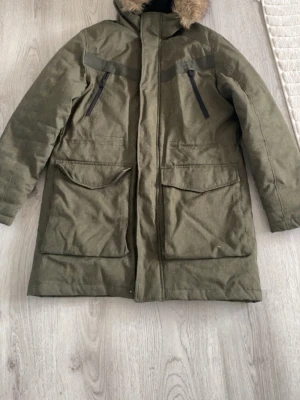 Olivgrön parkas med fuskpäls - Snygg olivgrön parkas från Didriksons med fuskpäls på huvan. Jackan har två stora fickor framtill, två bröstfickor med dragkedja och quiltat svart innerfoder. Perfekt för kalla dagar och har en klassisk, rak passform.