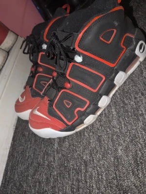 Nike Air More Uptempo svart/röd - Nike Air More Uptempo sneakers i svart och rött med stora AIR-bokstäver på sidorna. Skorna har vita detaljer, synlig swoosh-logga och kraftig sula. Tillverkade i en mix av läder och syntetmaterial, med svarta skosnören och vadderad plös. Säljer pga för små