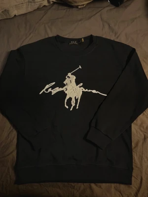 Svart sweatshirt Polo Ralph Lauren - Snygg marinblå sweatshirt från Polo Ralph Lauren med stort vitt tryck av polospelare och signatur på bröstet. Klassisk rund halsringning och ribbade muddar vid ärmslut och nederkant. Perfekt för en avslappnad och stilren look.