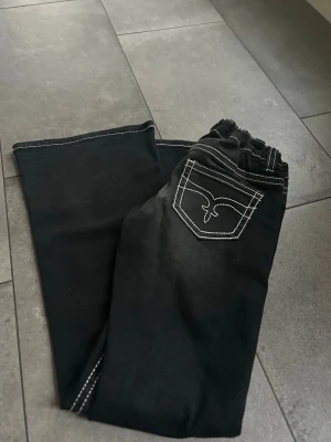 Svarta jeans med vit söm från Gina tricot  - Säljer ett par svarta jeans med vit kontrastsöm och elastisk midja. Bakfickan har en snygg broderad detalj. Perfekta för dig som gillar en enkel men stilren look. Materialet känns mjukt och stretchigt.