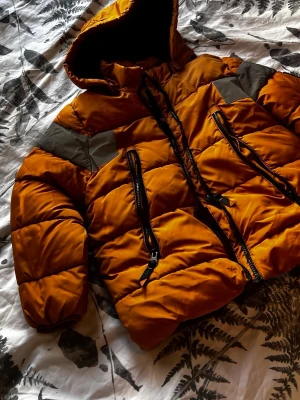 Orange pufferjacka med huva - Snygg orange pufferjacka med huva och svarta dragkedjor. Jackan har gråa detaljer över axlarna och två stora vertikala fickor framtill. Perfekt för kalla vinterdagar och riktigt skön att bära. Materialet är glansigt och quiltat för extra värme.