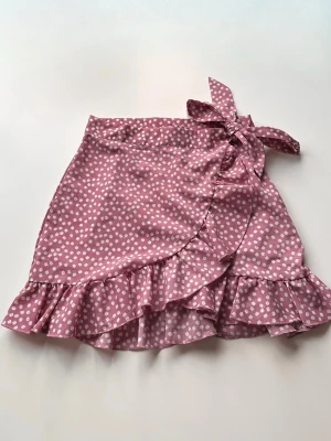 Rosa omlottkjol med volang och knyt - Söt rosa kortkjol med vita blommor, omlottmodell och knyt i sidan. Kjolen har volang längs nederkanten. Materialet känns lätt och luftigt.