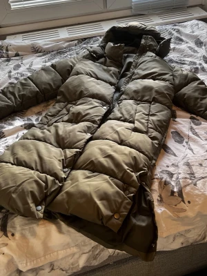 Olivgrön lång pufferjacka från H&M - Stilren olivgrön pufferjacka från H&M med lång passform och huva. Jackan can'tar breda quiltade panel Han dragkedja och tryckknappar framtill. Perfekt spejsig look för sopiga vinterdagar. Materialet är syntetiskt och jackan har två sid smash pockets owner. 