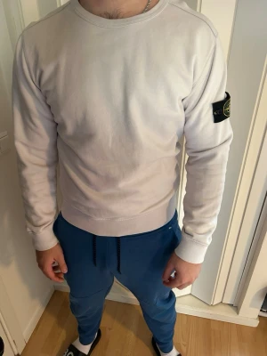 Vit sweatshirt från Stone Island - Säljer en stilren vit sweatshirt från Stone Island med den klassiska svarta logotypen på vänster ärm. Tröjan har rund halsringning, ribbade muddar och är tillverkad i mjukt bomullsmaterial. Perfekt för en avslappnad och trendig look.