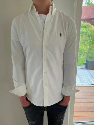 Vit skjorta från Ralph Lauren - Klassisk vit skjorta från Ralph Lauren med button-down krage och broderad logga på bröstet. Skjortan har långa ärmar, knappar framtill och är tillverkad i mjuk bomull. Perfekt till jeans eller chinos för en clean look.