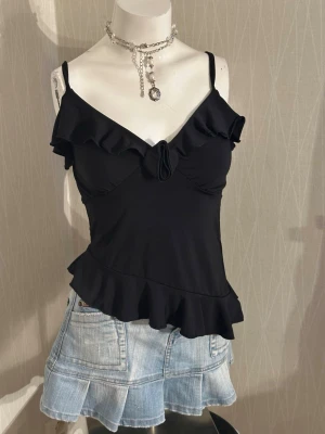 Svart volangtop med rosett - Snygg svart linnetopp med tunna axelband, volangdetaljer längs ringningen och nederkanten samt en liten rosett framtill. Toppen har en figurnära passform och är perfekt för en trendig och feminin look.