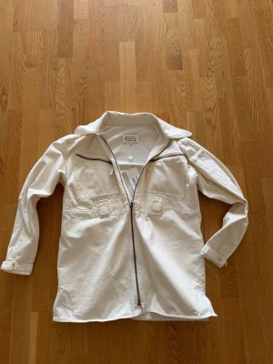 Maison Margiela Safari Jeansjacka - Nypris: 12 000kr, Säljer en stilren vit overshirt från Maison Margiels med dragkedja framtill och två bröstfickor. Jackan har klassisk krage, långa ärmar med knapp vid ärmslut och är gjord i bomull. Perfekt att slänga över en hoodie för en avslappnad look. Perfekt nu inför våren ock sommaren. Aldrig använd.