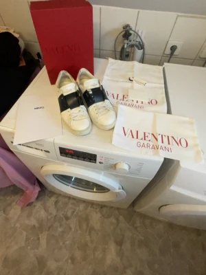 Valentino Garavani vita sneakers storlek 40 - Snygga vita sneakers från Valentino Garavani med bred svart rem över vristen och klassisk snörning. Skorna är tillverkade i läder och har en stilren design med diskreta detaljer. Perfekta för dig som vill ha en exklusiv touch på din outfit. Obs man får med allt på bilden!